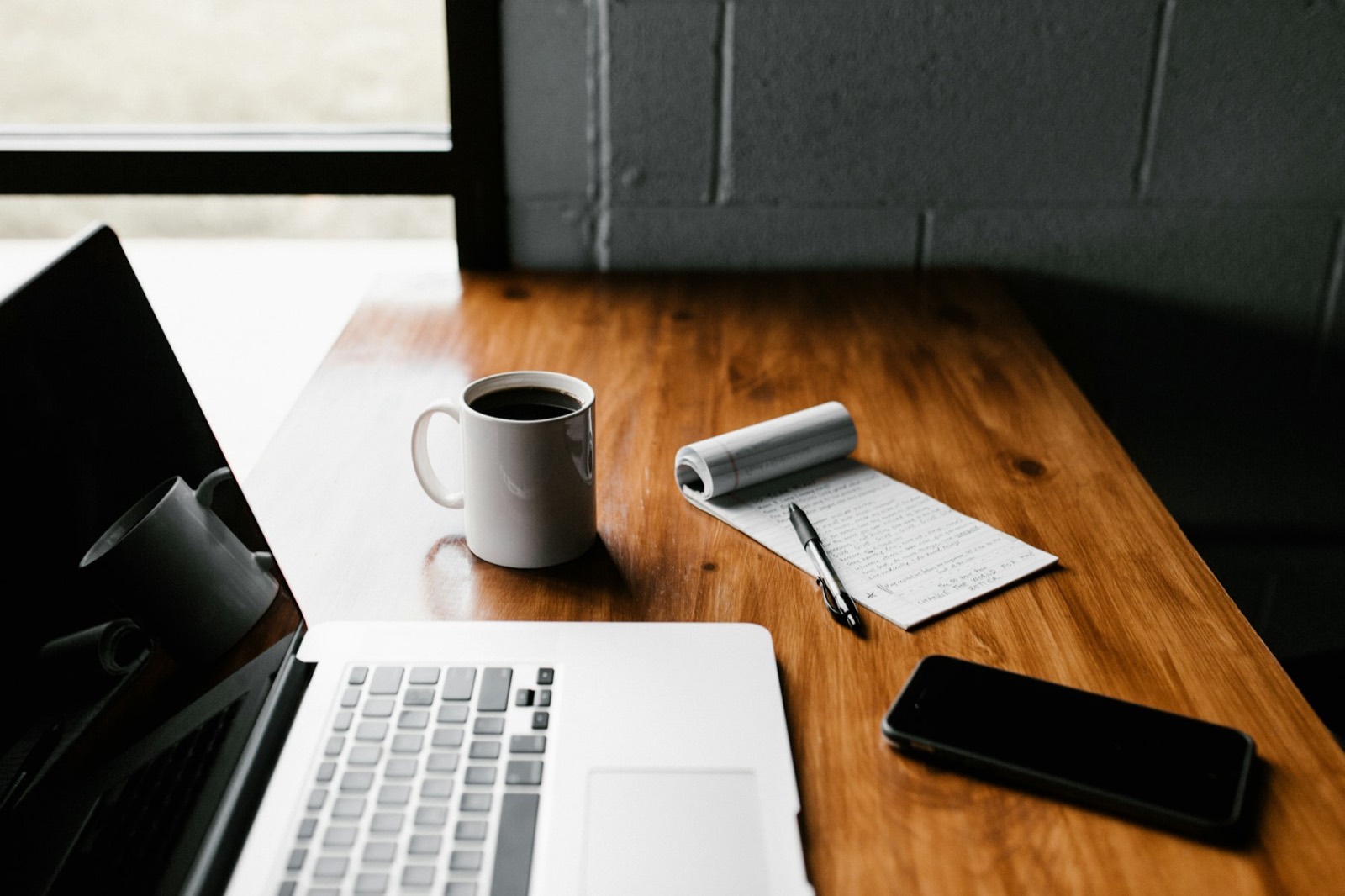 A tidy work desk in warm window light — laptop, coffee and a handwritten notepad